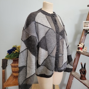 May include: A gray patchwork cardigan sweater with a round neckline and button-up front. The sweater features various shades of gray in geometric shapes. The sleeves are long and the overall style is oversized.