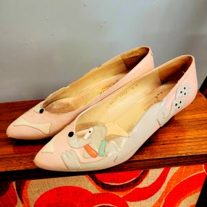 May include: A pair of pink leather flats with cartoon dog designs. The shoes have a pointed toe and a low heel. The dog design features a black eye, a beige body, and an orange and green tongue. The shoes are size 6M.