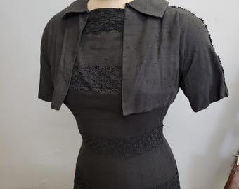 Puede incluir: Un conjunto vintage de vestido negro y chaqueta bolero. El vestido presenta una silueta ajustada con detalles de encaje alrededor del corpiño y la falda. La chaqueta bolero tiene mangas cortas y un cuello con solapas, creando un look clásico y elegante. El conjunto se exhibe en un maniquí.