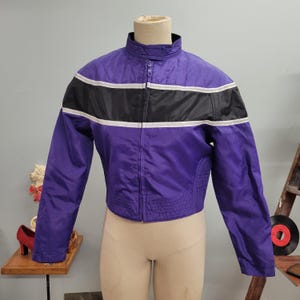 May include: A vintage purple jacket with a black and white stripe across the chest. The jacket has a stand-up collar, a full zipper, and a cropped waist. The jacket is displayed on a mannequin and appears to be made of a nylon or similar material.