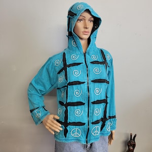 May include: A bright turquoise zip-up hoodie featuring a grid of black fabric strips and white embroidered spiral patterns. The garment includes a hood and long sleeves, with white embroidered peace sign symbols on the lower front and cuffs.