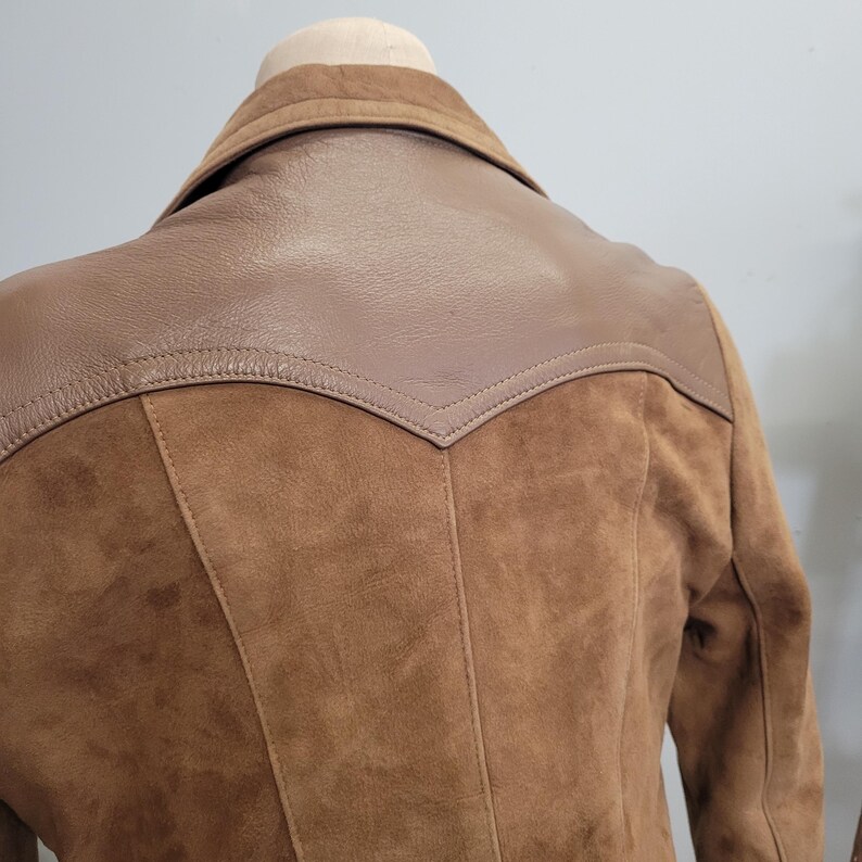 May include: Brown suede jacket with a leather yoke on the back. The jacket has a collar and long sleeves.