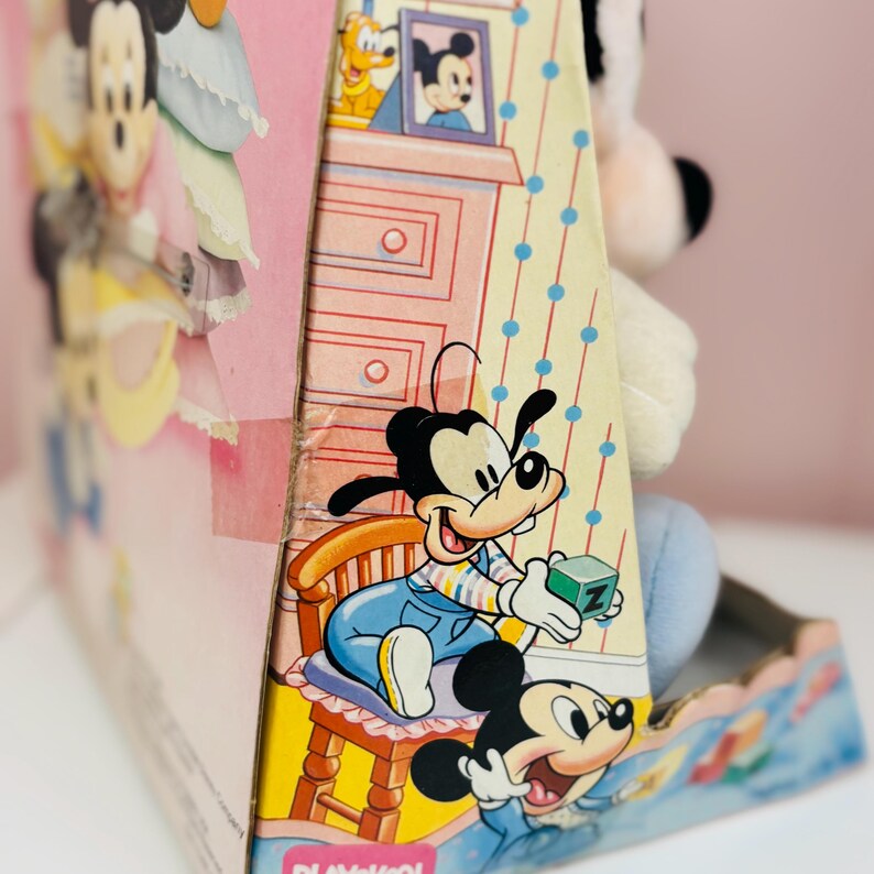 May include: A vintage Mickey Mouse plush toy peeking out of its original box. The box features colorful illustrations of Mickey Mouse and Goofy in a nursery setting. The box is pink and yellow with a blue polka dot pattern.