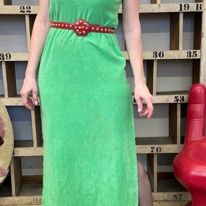 Puede incluir: Un vestido maxi verde con capucha y un cintur&oacute;n rojo con tachuelas. El vestido est&aacute; hecho de un material suave de felpa. La modelo lleva tacones negros y medias negras.