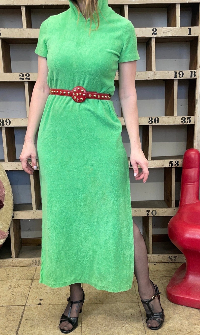 Puede incluir: Un vestido maxi verde con capucha y un cintur&oacute;n rojo con tachuelas. El vestido est&aacute; hecho de un material suave de felpa. La modelo lleva tacones negros y medias negras.