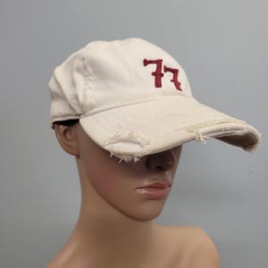 May include: Cream-coloured baseball cap with a distressed peak. The cap displays the number "77" embroidered in maroon on the front. The hat is made of a soft, worn fabric, suggesting a vintage or casual style.
