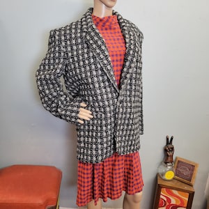 May include: A black and white patterned blazer with a notched collar, worn over a red and purple plaid dress. The blazer has a textured weave and padded shoulders. The dress has a high neckline and long sleeves.