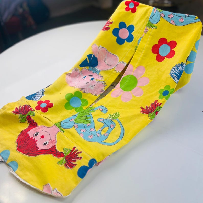 May include: Folded yellow fabric with cartoon characters and floral patterns. The design includes a red-haired doll, pink animals, and blue and pink flowers. The fabric is likely vintage and suitable for crafting or decorative purposes.