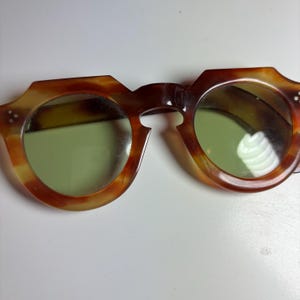 May include: Tortoiseshell-patterned sunglasses with a distinct geometric frame shape and round lenses. The frames feature a brown and amber mottled design with three small silver-toned rivets on each outer corner. The lenses have a light green tint.