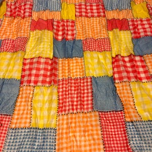 May include: A patchwork quilt featuring a repeating grid of square fabric blocks in vibrant colors. The pattern consists of red, orange, yellow, and blue gingham checks, joined together with visible black cross-stitch embroidery detailing.