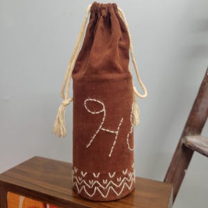 Pode incluir: Uma bolsa para garrafa de vinho em veludo cotelê castanho com fecho de cordão e corda creme. A bolsa apresenta detalhes bordados brancos, incluindo a letra "H" e um padrão decorativo na base. Projetada para uma garrafa de vinho padrão.