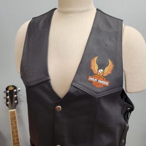 May include: Black leather vest with a Harley Davidson Cycles patch featuring an eagle. The vest has a button-up front and a classic biker style. The vest is displayed on a mannequin.