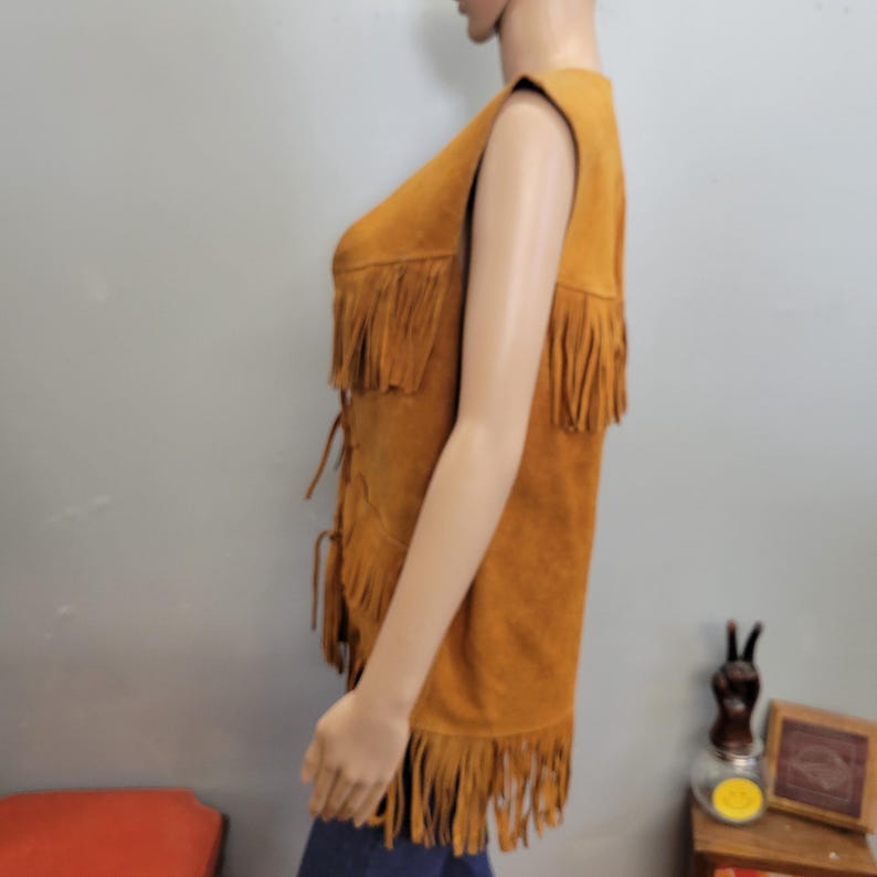 May include: A tan suede vest with fringe detailing. The sleeveless vest features multiple rows of fringe across the chest and bottom hem. It has a lace-up closure down the front. The vest is displayed on a mannequin.