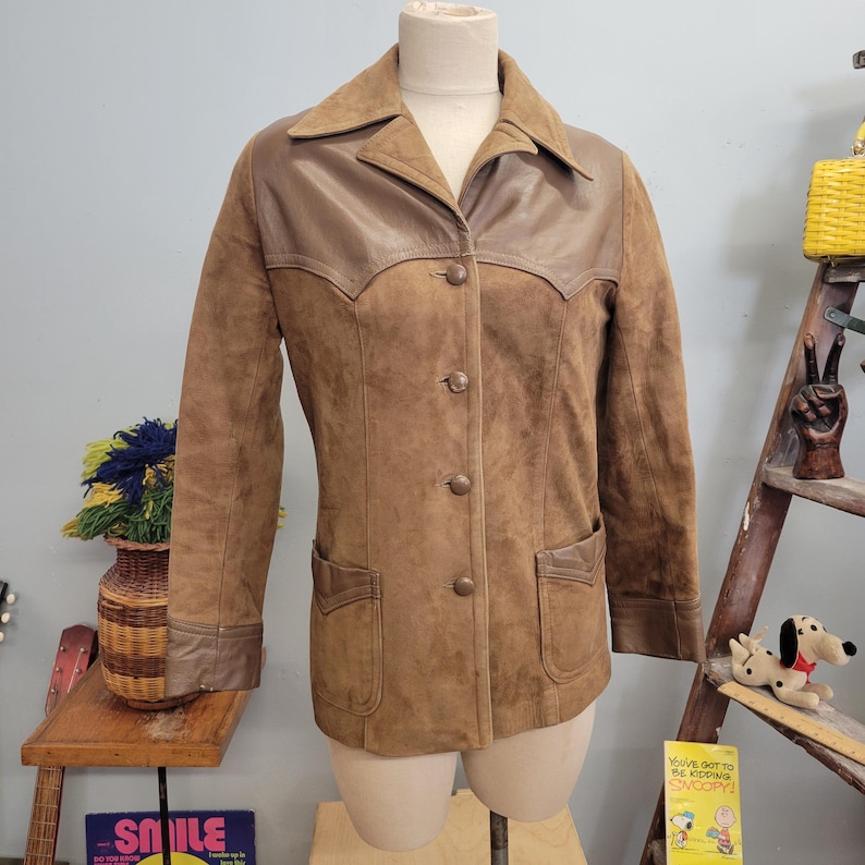May include: A brown suede jacket with a brown leather collar and pockets. The jacket has a button-up front and long sleeves.