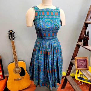 May include: A sleeveless dress featuring a vibrant blue base with a repeating pattern of green and brown geometric blocks and floral motifs. The dress has a fitted bodice, a defined waistline, and a full, pleated skirt that falls to the knees.