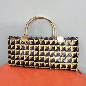 Puede incluir: Un bolso negro rectangular con un patrón geométrico en amarillo y dorado. El bolso tiene un asa y un cierre de metal dorado. El bolso está sobre una superficie naranja.