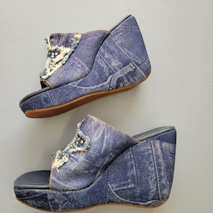 Puede incluir: Un par de sandalias de cuña vaqueras. Los zapatos de punta abierta tienen una parte superior de mezclilla azul con un acento decorativo en la pala. El tacón de cuña mide aproximadamente 10 cm de alto, con una suela marrón.