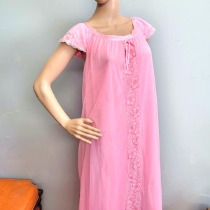 May include: A floor-length light pink nightgown featuring short flutter sleeves with delicate lace trim. The garment has a scoop neckline with a small tie detail and a vertical panel of floral lace embroidery running down the front center.