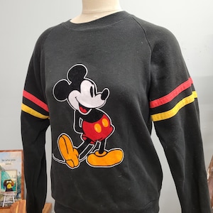 May include: Black sweatshirt featuring a large graphic of Mickey Mouse in his classic pose. The sleeves have red and yellow stripes. The sweatshirt is displayed on a mannequin.