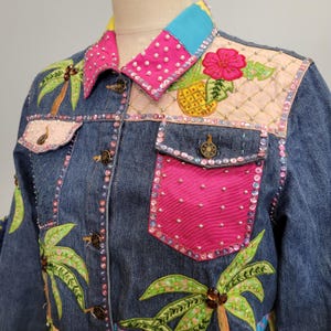 Könnte beinhalten: Jeansjacke mit farbenfrohen tropischen Stickereien. Zu den Merkmalen gehören ein rosa Kragen mit weißen Perlenakzenten, ein türkisfarbener Kragenakzent und eine rosa Tasche. Die Jacke ist mit Palmen, einer Ananas und einer Blume verziert.