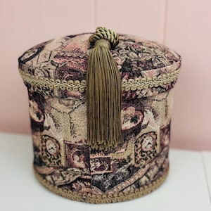 May include: A cylindrical fabric storage box featuring a tapestry-style pattern of clocks and portraits in shades of brown, black, and cream. The lid is adorned with a long, olive-green decorative tassel and gold braided trim along the edges.