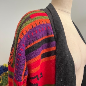 May include: A colorful, patterned cardigan sweater with a black collar. The sweater features a vibrant design with red, orange, purple, green, and black stripes and geometric shapes. The sweater is displayed on a mannequin.
