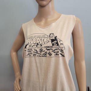 May include: A sleeveless beige tank top featuring a black graphic print. The design includes the text "I HELPED FINISH OFF UPPER DECK" above a cartoon character wearing a "KISS AT OFF" shirt, with a list of song titles including "GROUND ZERO" and "URBAN GUERILLAS."