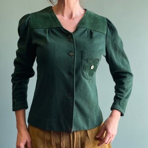May include: A dark green button-up jacket with a flower-shaped patch on the pocket. The jacket has long sleeves and a v-neck.