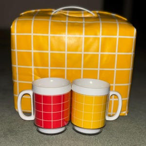 May include: Two ceramic mugs with white handles and rims, one red and one yellow, each with a grid pattern. A matching yellow bag with a white grid pattern and a handle is behind the mugs.