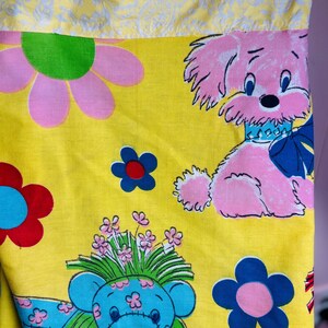 May include: Bright yellow fabric featuring a repeating pattern of pink flowers, blue flowers, and cartoon animals. The cartoon animals include a pink dog with a blue bow and a blue bear with a bouquet of flowers.