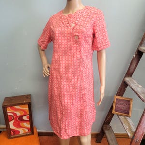 May include: A knee-length dress in a vibrant coral and white grid pattern. The garment features short sleeves, a round neckline, and a wrap-style front closure secured by two round buttons positioned on the upper right side of the bodice.