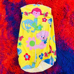 May include: A bright yellow garment with a retro design. It showcases a cartoon girl, a blue creature, and a pink dog with a blue collar. The design incorporates flowers and other colourful elements. The garment is suspended from a metal hanger.
