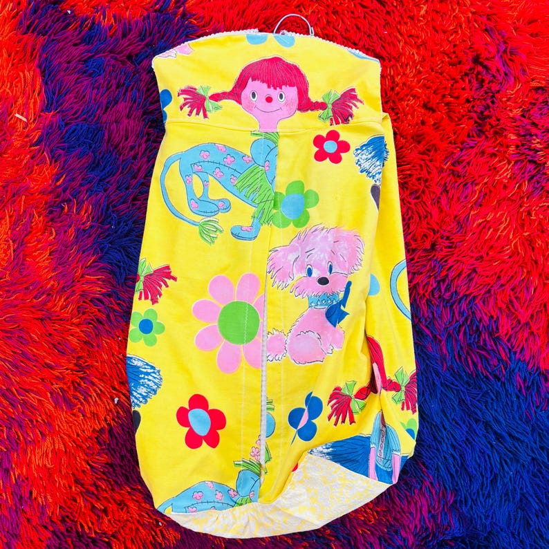 May include: A bright yellow garment with a retro design. It showcases a cartoon girl, a blue creature, and a pink dog with a blue collar. The design incorporates flowers and other colourful elements. The garment is suspended from a metal hanger.