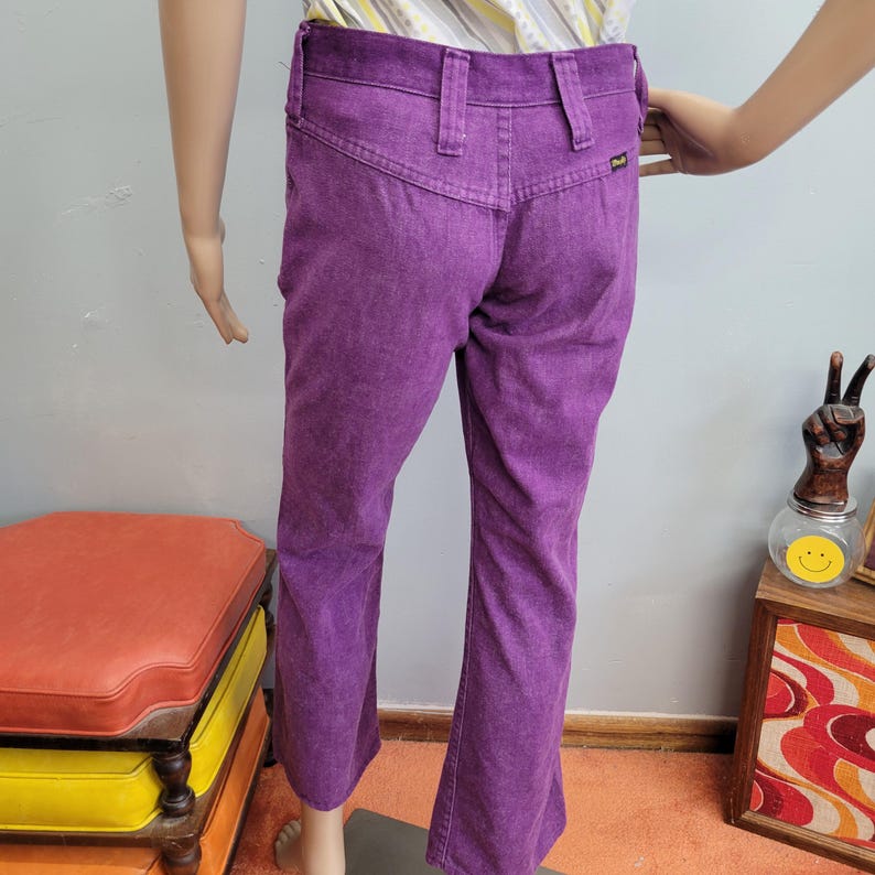 May include: A pair of vintage purple corduroy pants. The pants have a high waist, belt loops, and a flared leg. The back pockets are visible. The pants are displayed on a mannequin.