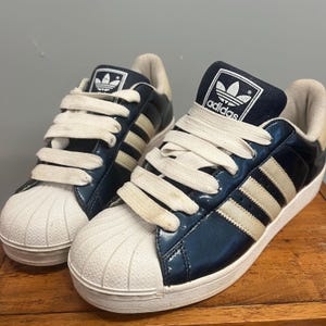 May include: A pair of navy blue and white Adidas Superstar sneakers. The shoes feature white laces, a white toe cap, and the iconic three stripes in off-white. The Adidas logo is visible on the tongue.