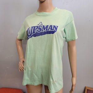 May include: A light green t-shirt with the word "UPSMAN" in blue lettering. The shirt has short sleeves and a crew neck. The shirt is displayed on a mannequin.