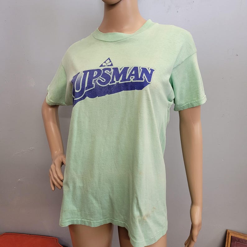May include: A light green t-shirt with the word "UPSMAN" in blue lettering. The shirt has short sleeves and a crew neck. The shirt is displayed on a mannequin.