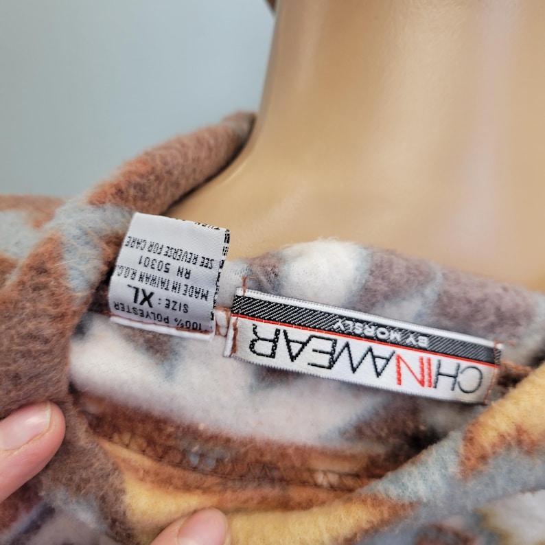 K&ouml;nnte beinhalten: Nahaufnahme eines Fleece-Kleidungsst&uuml;cks mit einem Muster in Braun-, Wei&szlig;- und Blaut&ouml;nen. Das Etikett lautet "CHINAWEAR BY MORSLEY" und weist die Gr&ouml;&szlig;e XL aus. Der Stoff besteht aus 100 % Polyester.