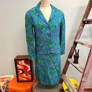 May include: A vintage two-piece suit featuring a blazer and skirt. The suit is patterned with a blue and green paisley design. The blazer has a notched collar and button closure. The skirt is knee-length. The suit is displayed on a mannequin.