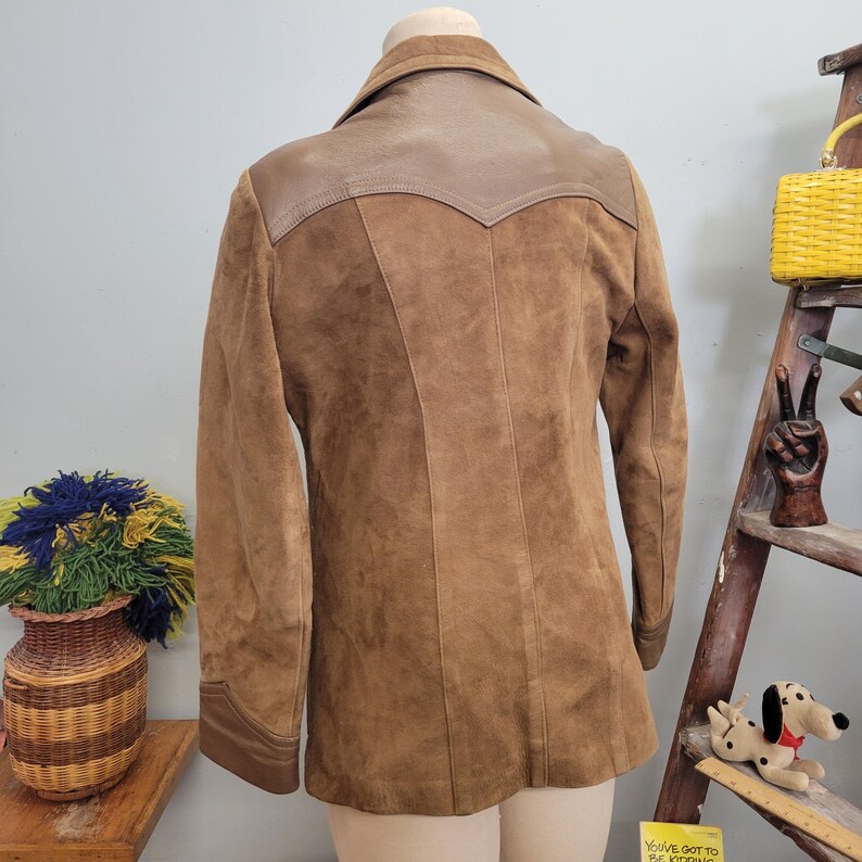 May include: A brown suede jacket with a brown leather yoke. The jacket has a long sleeve and a collar.