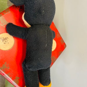 May include: A vintage black plush toy with white face and ears, and yellow feet, is leaning against a red, square-shaped object with gold-colored circles. The object has a distressed, aged appearance.