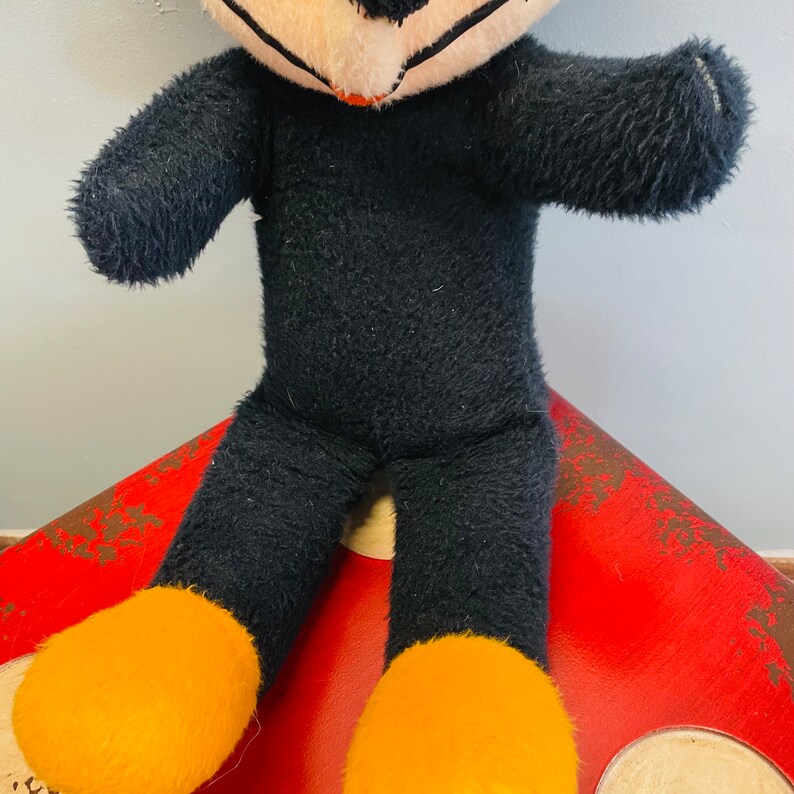 May include: A vintage plush toy cat with black fur, a white face, and orange feet. The cat is sitting on a red and brown surface. The toy has a stitched mouth and black eyes.