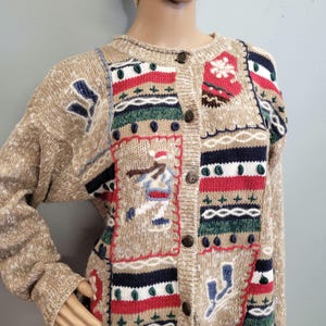 Pode incluir: Cardigã bege com botões e design de inverno. O suéter de malha apresenta patches bordados com esquiadores, flocos de neve e padrões geométricos em vermelho, azul, verde e branco. Gola redonda e mangas compridas.