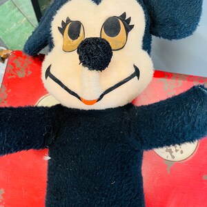 May include: Vintage plush toy featuring a black and cream-colored character with large eyes and a black nose. The toy has black arms and ears, and a smiling mouth. The toy is set against a red background.