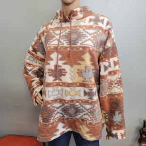 K&ouml;nnte beinhalten: Ein Fleece-Pullover mit hohem Kragen und Kordelzug. Der Pullover hat ein vom S&uuml;dwesten inspiriertes Muster in Braun-, Creme-, Grau- und Gelbt&ouml;nen. Das Design umfasst geometrische Formen und stilisierte Vogelmotive. Der Pullover wird auf einer Schaufensterpuppe pr&auml;sentiert.
