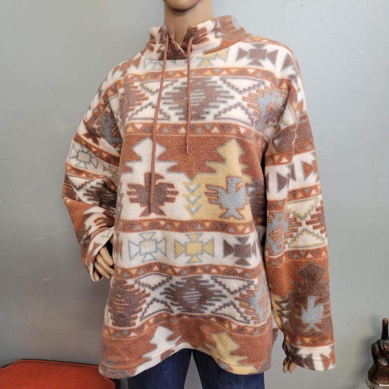 K&ouml;nnte beinhalten: Ein Fleece-Pullover mit hohem Kragen und Kordelzug. Der Pullover hat ein vom S&uuml;dwesten inspiriertes Muster in Braun-, Creme-, Grau- und Gelbt&ouml;nen. Das Design umfasst geometrische Formen und stilisierte Vogelmotive. Der Pullover wird auf einer Schaufensterpuppe pr&auml;sentiert.
