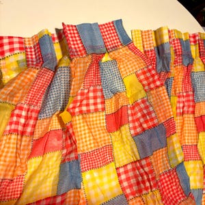 May include: A patchwork fabric with a variety of gingham patterns in red, yellow, orange, and blue. The fabric is gathered at the top, creating a ruffled effect. The squares are stitched together with a contrasting thread.