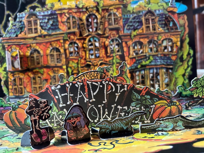 以下が含まれることがあります： お化け屋敷、カボチャ、切り絵のフィギュアが特徴の、ポップアップハロウィーンデコレーション。看板には「Happy Halloween」と白文字で黒い背景に書かれています。家はオレンジと茶色です。