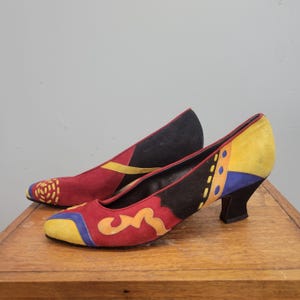 May include: A pair of low-heeled shoes in a vibrant colour palette. The shoes combine red, black, yellow, and blue suede with decorative patterns. The heels are black and the shoes are on a wooden surface.