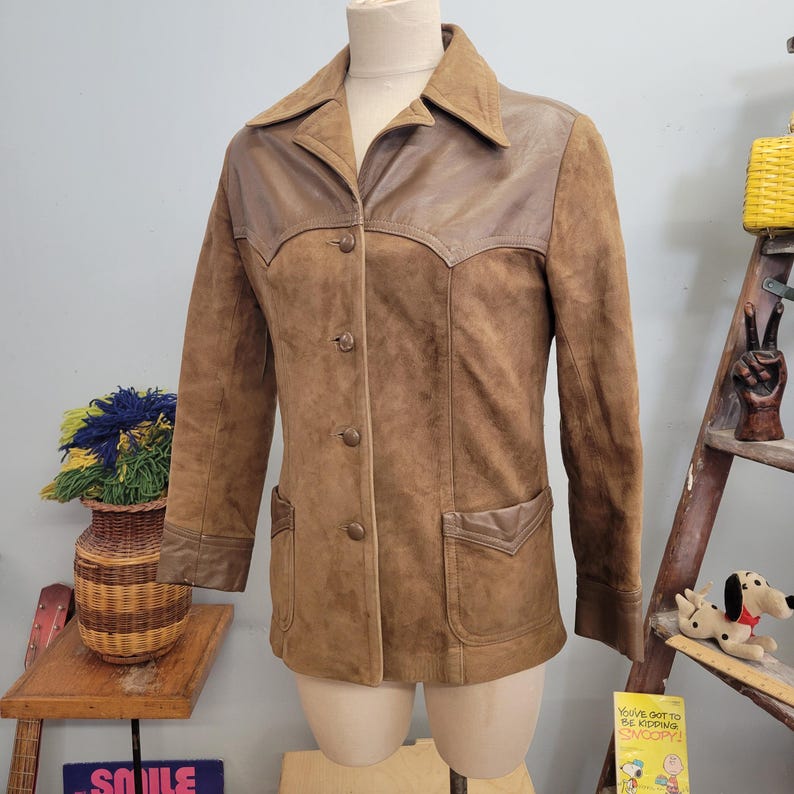 May include: A brown suede jacket with a brown leather collar and pockets. The jacket has a button-up front and long sleeves.